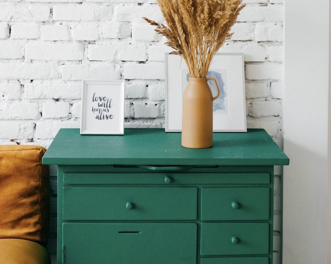 Green table full of drawers with a vase of wheat and two pictures aside a brown couch 